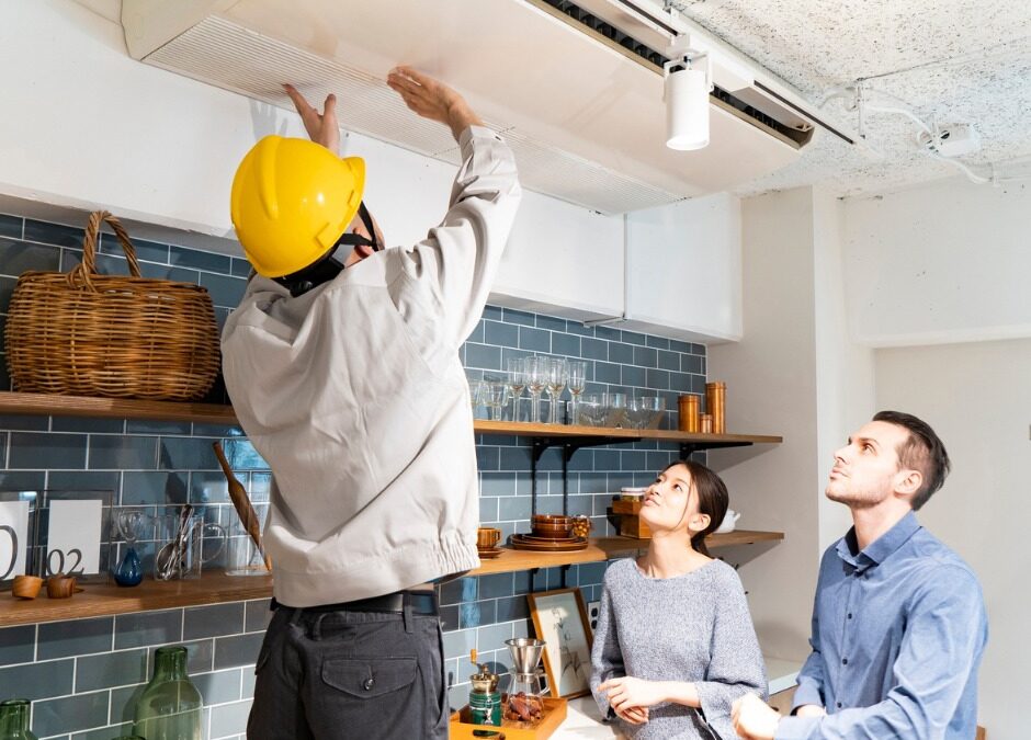 A technician in a yellow hard hat inspects a ceiling-mounted air conditioning unit in a modern kitchen while a couple watches from below